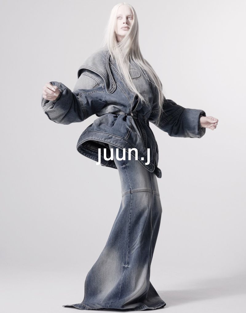 A lone, androgynous model stands on a seamless backdrop, showcasing a blue denim jacket and wide-leg denim trousers in a fashion editorial.