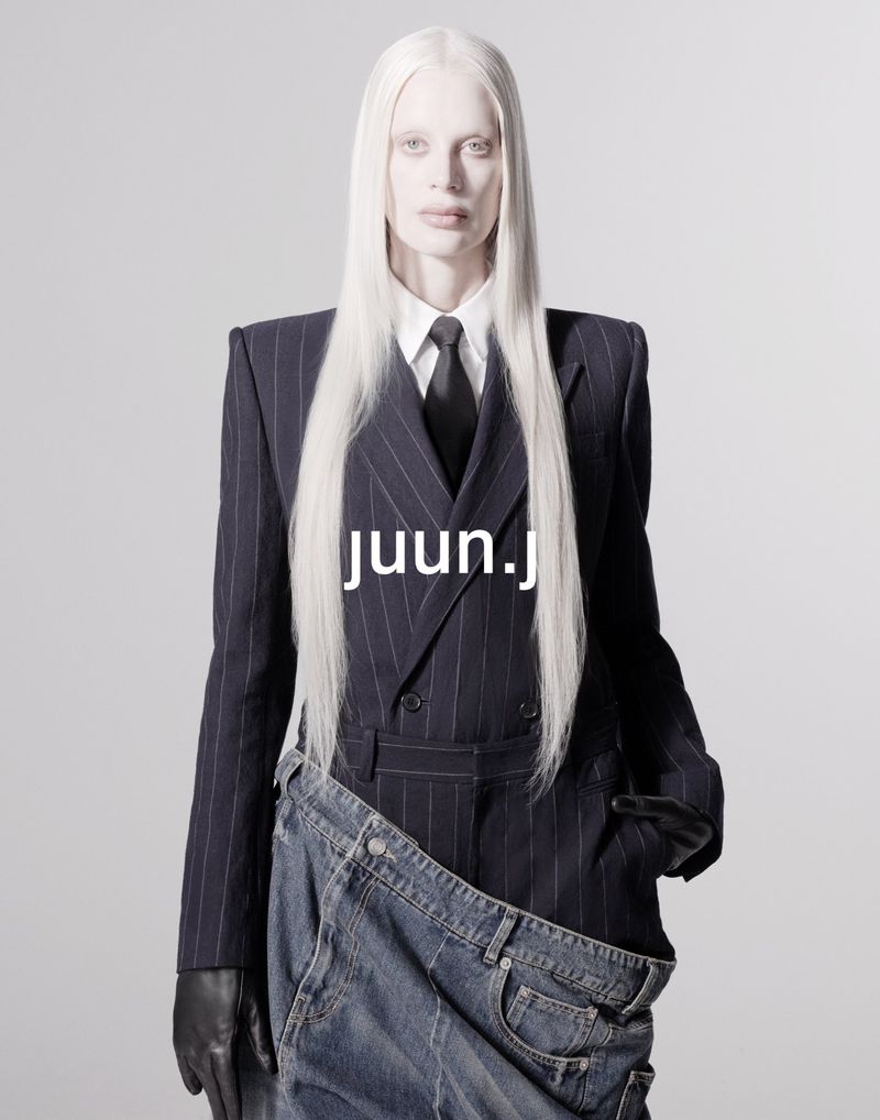 Woman with albino skin and long white hair wearing a pinstripe suit jacket and tie with denim garment, posing against a gray backdrop.