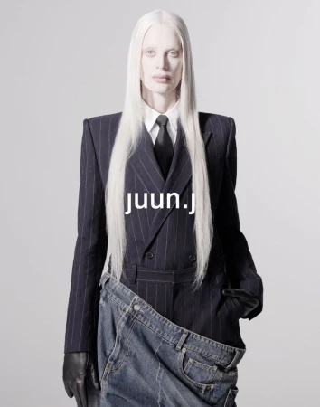 Woman with albino skin and long white hair wearing a pinstripe suit jacket and tie with denim garment, posing against a gray backdrop.