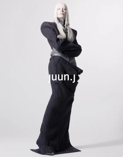 Model Kristen McMenamy wearing an exaggerated shoulder pinstripe cropped blazer and floor-length trousers against a white background.