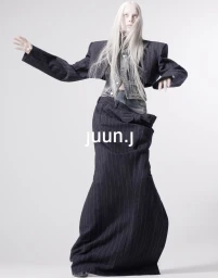 Model Kristen McMenamy in structured pinstripe blazer and long skirt against a plain white studio backdrop.