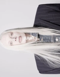 Kristen McMenamy with long platinum blonde hair wearing a denim jacket under a dark pinstripe blazer against a white backdrop.
