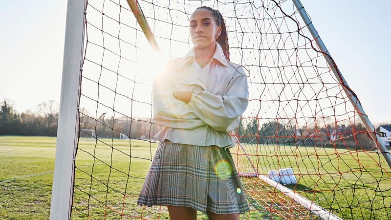 Naomi Girma standing inside a soccer goal net wearing a light beige jacket and plaid pleated skirt in bright sunlight.