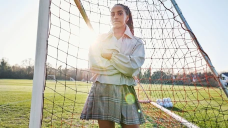 Woman standing with arms crossed in front of a soccer goal net wearing a light jacket and plaid skirt.