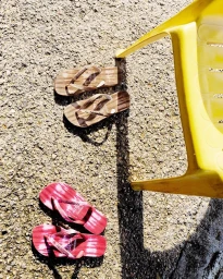 Two pairs of flip flops and a yellow plastic chair cast shadows on a textured outdoor ground surface.