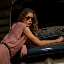 Model Hazel Jane wearing dark aviator sunglasses and a mauve pleated dress leaning on a vintage car for Brunello Cucinelli.