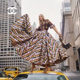 Model in a voluminous plaid dress stands barefoot on top of a yellow taxi cab on a city street for Chanel.
