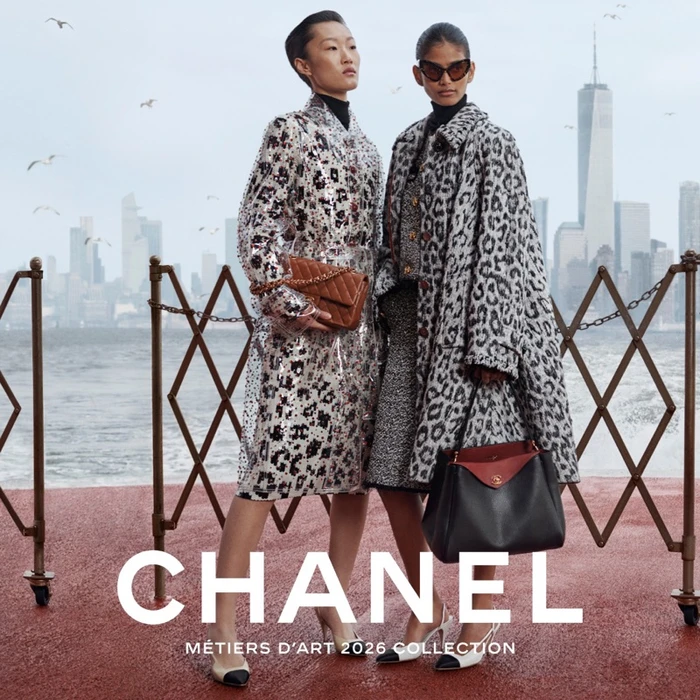 Two female models in Chanel coats posing by a waterfront railing with a city skyline in the background.