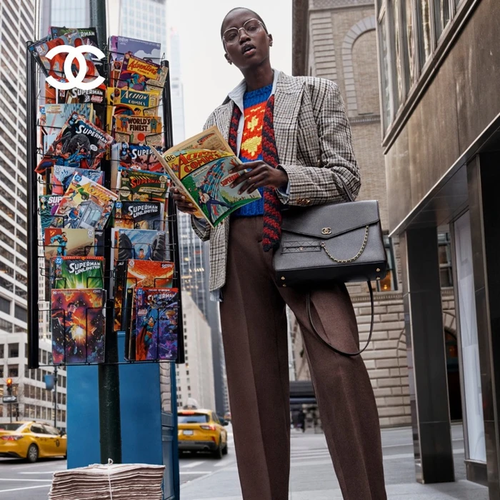 Model wearing a plaid blazer and brown trousers reads a comic book next to a newsstand on a city street.