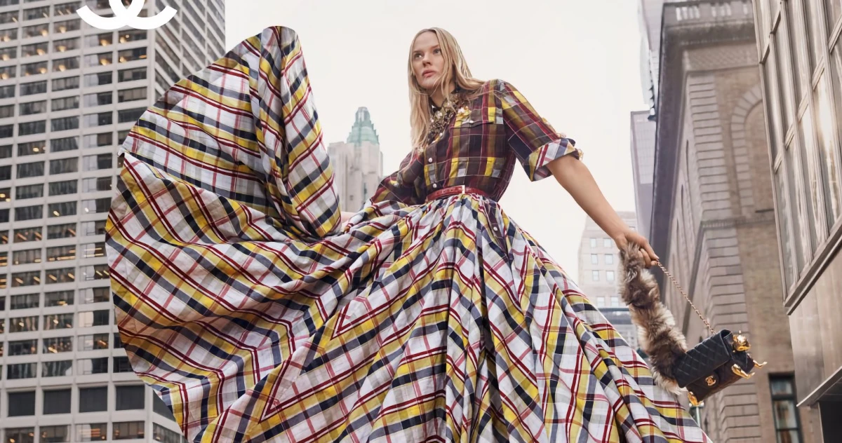 Model in a plaid shirt and full pleated skirt holding a quilted handbag against a city skyline