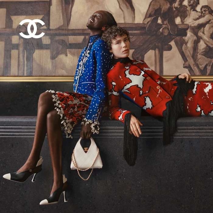 Two models posing together against a dark stone ledge with a large painting background for Chanel Pre-Fall 2026.