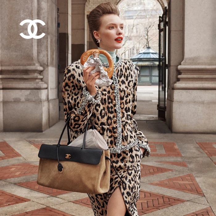 Model wearing a leopard print tweed jacket and skirt holding a pretzel and a black and tan handbag outdoors