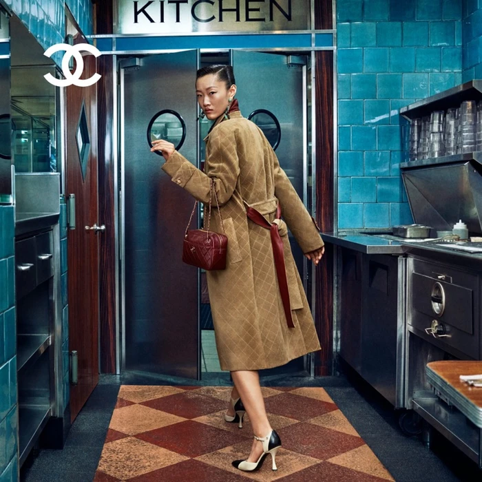 Model in quilted brown coat holding a burgundy handbag walking through a commercial kitchen with blue tiled walls