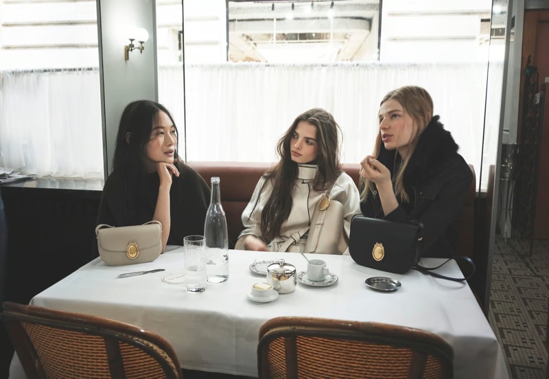 Three women seated at a restaurant table with white tablecloth, displaying luxury leather handbags with gold hardware.