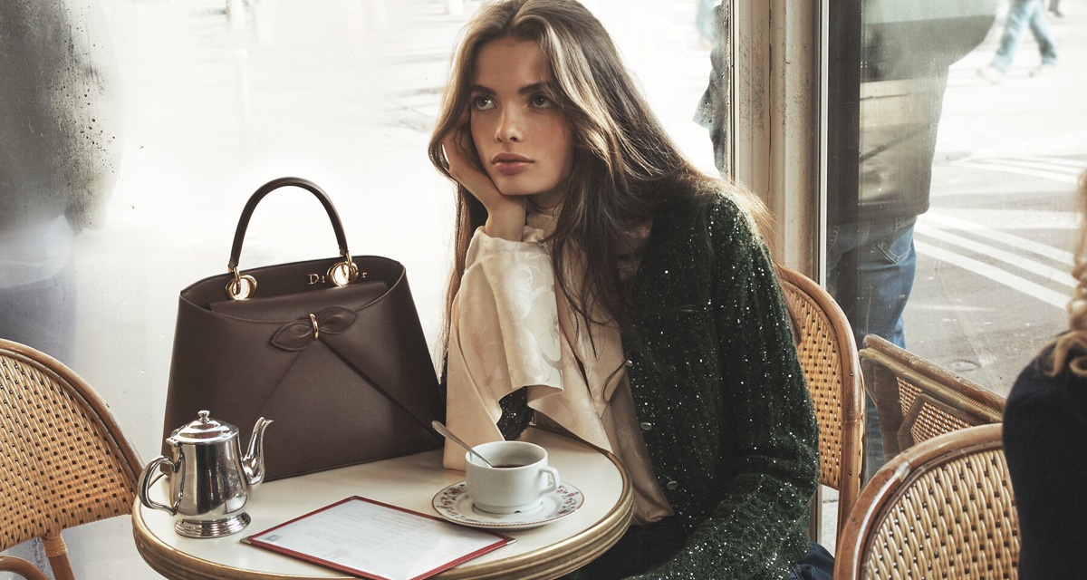 A woman in a green sequin cardigan resting her chin on her hand beside a brown leather handbag at a cafe table.