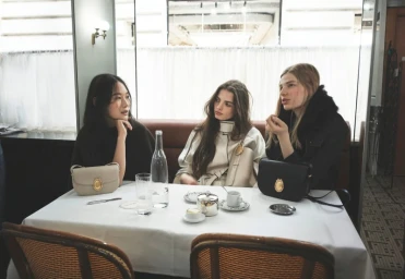 Three female models seated at a cafe table with handbags, photographed by David Sims for Dior Pre-Fall 2026.