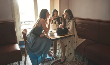Three female models seated together inside a café, wearing designer dresses and holding handbags.