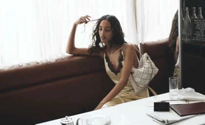 Model seated on a brown leather banquette in a cafe wearing a pale yellow silk slip dress with black lace trim.