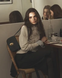 Model in cream blouse with bow detailing seated at cafe table wearing dark skirt and shoulder bag, photographed by David Sims for Dior
