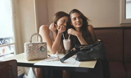 Two women sitting together at a cafe table with handbags for the Dior Pre-Fall 2026 campaign