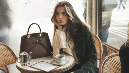 Model seated at a café table with a dark brown leather handbag, teapot, and cup, wearing a dark green textured jacket.