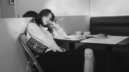 Model seated in a cane chair at a cafe table wearing a patterned shoulder bag for Dior pre-fall 2026 campaign.
