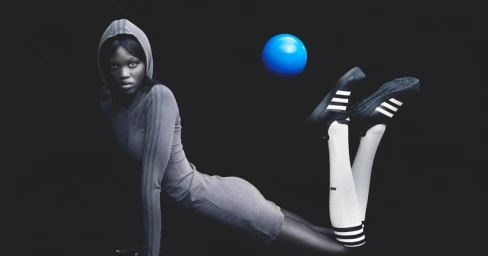 Model in grey hooded dress and white striped socks posing with a blue sphere against a dark background for Adidas.