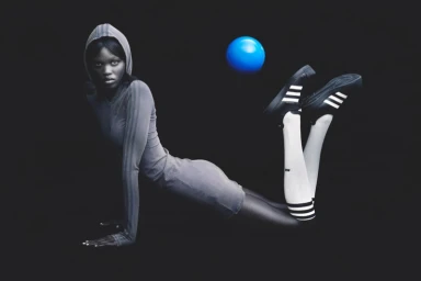 Model in grey hooded athletic dress and striped socks with sneakers posing against a black background with a blue ball