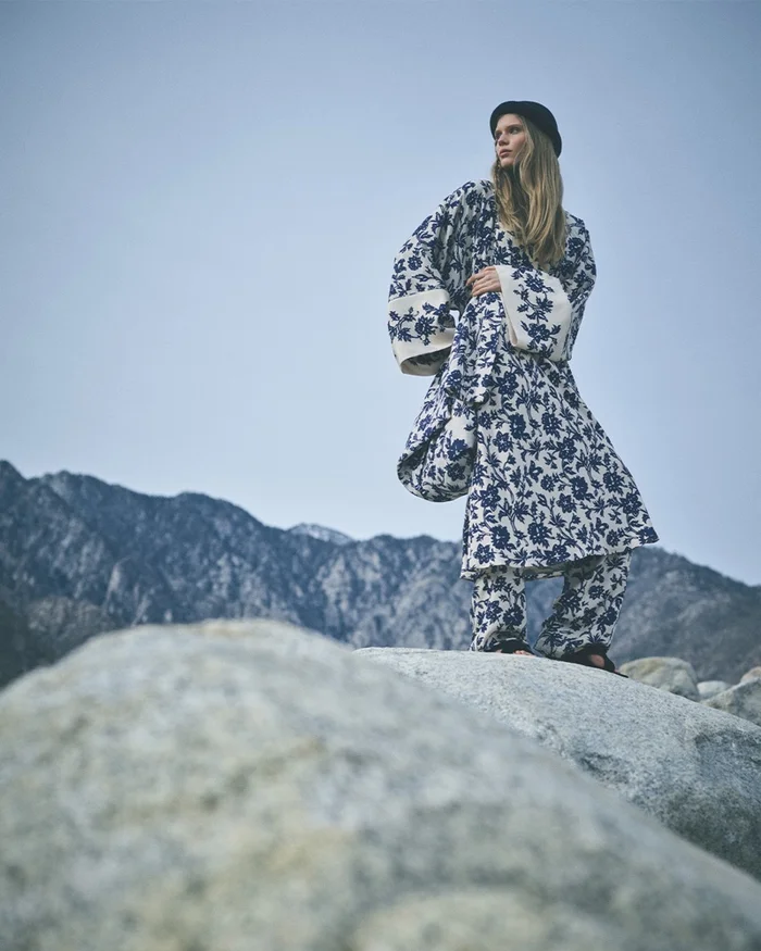 Model wearing a blue and white patterned outfit standing on a large rock with mountains and clear sky behind