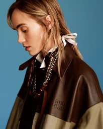 Close-up portrait of a woman in a brown leather jacket and white collar against a solid blue background