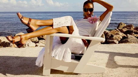 Model relaxing in a white wooden chair by the ocean, wearing sunglasses, a white dress, and brown sandals.