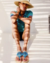 Model wearing a straw hat and striped top sitting on a light surface with striped shadows cast across her body.