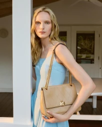 Model Alex Consani wearing a light blue dress holding a tan shoulder bag leaning against a white pillar