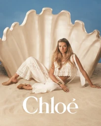 Model Apple Martin seated on sand in front of a giant seashell prop, wearing a white eyelet crop top and matching trousers for Chloé.