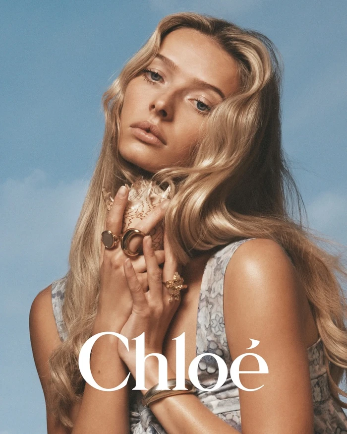 Model with long blonde hair holding a seashell wearing gold rings against a blue sky background for Chloé