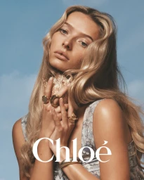 Model with long blonde hair holding a seashell wearing gold rings against a blue sky background for Chloé