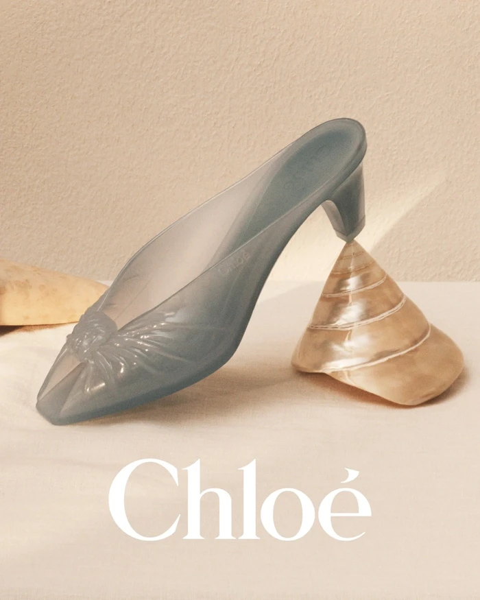 Translucent blue mule shoe balanced on a polished seashell against a neutral textured background with the Chloe logo below.