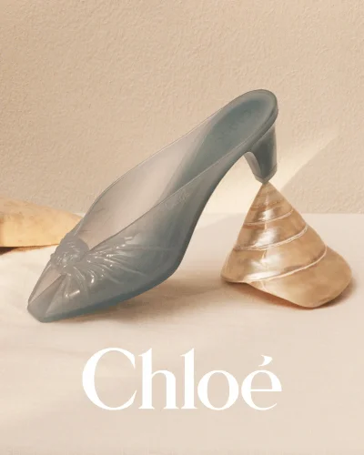Translucent blue mule shoe balanced on a polished seashell against a neutral textured background with the Chloe logo below.