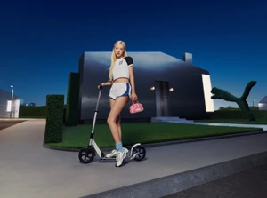 ROSÉ holding a pink handbag while riding a kick scooter in front of a dark modern house at dusk for Puma