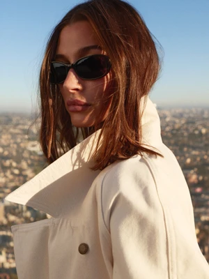 Hailey Bieber wearing dark sunglasses and a light coat against an urban rooftop backdrop