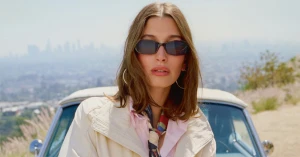 Hailey Bieber wearing sunglasses and a light jacket in front of a car with a city skyline in the background