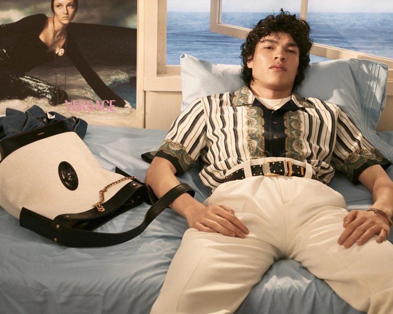 Model Alvise Candida lounging on a bed in a patterned shirt and white trousers next to a canvas tote for Versace.