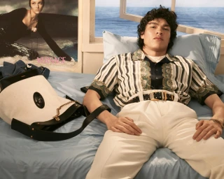 Model lying on a bed wearing a patterned shirt and white trousers next to a white and black Versace handbag.