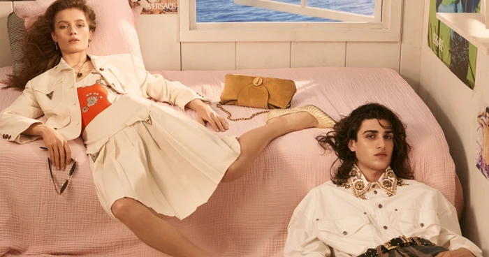 Model reclining on a pink bed in a white jacket and skirt alongside another model in a white shirt, photographed by Steven Meisel for Versace.