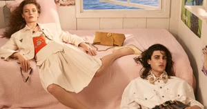 Model reclining on a pink bed in a white jacket and skirt alongside another model in a white shirt, photographed by Steven Meisel for Versace.