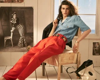 Model seated backwards on wooden chair wearing a light blue shirt and red trousers against a wood-panelled wall with posters.