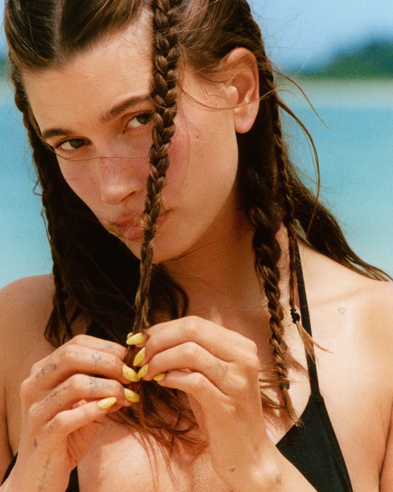 Close-up of a young woman on a beach, featuring braided hair, yellow nail polish, and a black bikini top against a blurred background of the sea and sky.