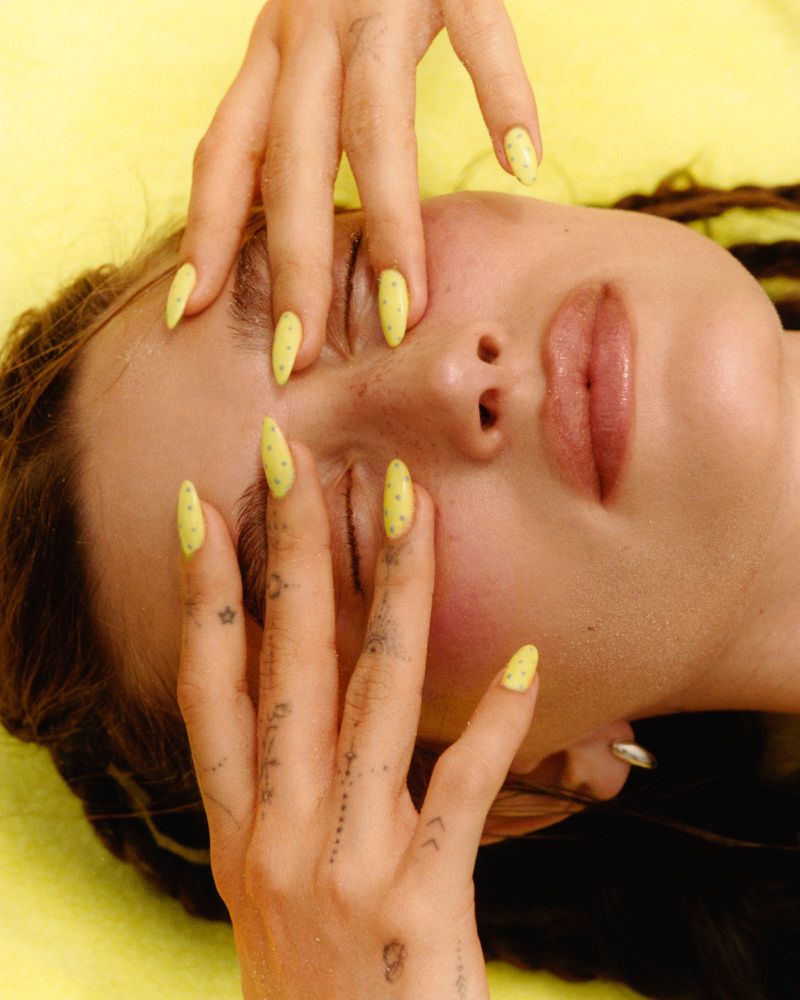 Eye-level close-up of a fair-skinned woman lying down with her hands over her face, manicured nails painted pastel yellow with dots.