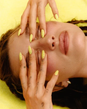 Hailey Bieber with closed eyes framing her face with yellow stiletto nails and finger tattoos against a yellow background.
