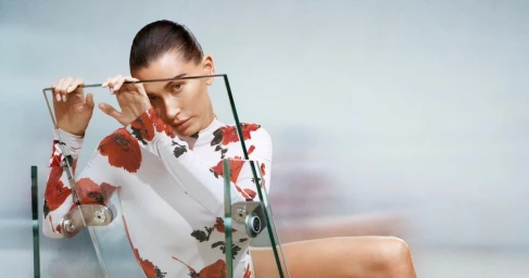 Hailey Bieber wearing a white and red floral top behind a glass panel for Alaia